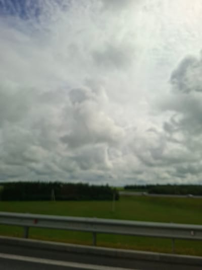 The image captures a blurry view of a cloudy sky over a green landscape, with a guardrail in the foreground. The scene appears to be a roadside environment, possibly near a highway, as suggested by the presence of the guardrail and the flat, open terrain. There are no discernible people, specific landmarks, or buildings visible to identify the precise location within Pokrovka, Russia, or any other city. The overall impression is one of a nondescript, overcast day in a rural or suburban area.