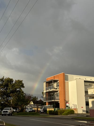 A landscape-oriented shot shows a partial rainbow in a cloudy sky, with buildings, trees, power lines, and a road visible. The building on the right has a brick facade on the top story, transitioning to white on the levels above. The street includes parked white vehicles. The scene is likely in Bassendean, Australia.