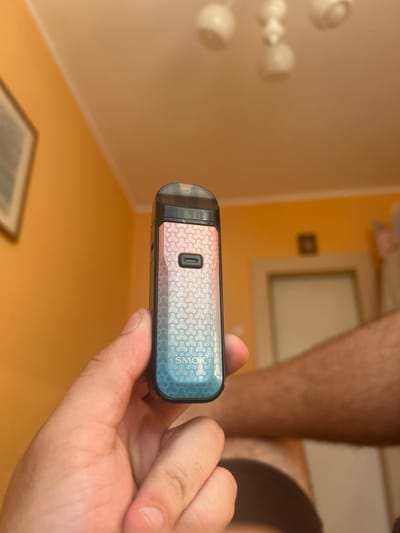 A close-up shot shows a person's hand holding a vape device. The vape device is black with a colorful gradient pattern featuring a geometric design and the "SMOK" logo. The background is out of focus, but appears to be an indoor setting with an orange wall, a doorway, and a ceiling fixture with multiple lights. The overall lighting suggests a dimly lit room. There is no visible information to determine the location within Tomaszów Mazowiecki, Poland.