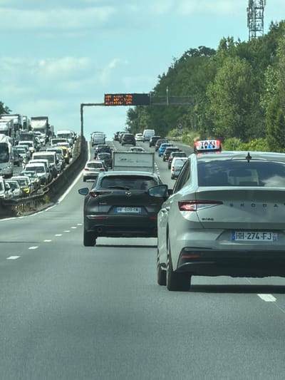 The image depicts a traffic jam on a highway in Savigny-sur-Orge, France. Cars are lined up bumper-to-bumper, indicating a significant delay. The scene is dominated by vehicles, including cars, trucks, and what appears to be a taxi with illuminated signs. The sky is visible and appears bright, suggesting it is daytime, possibly late morning or afternoon, and the weather seems to be clear.
On the side of the road, there is a traffic sign displaying amber text that is difficult to decipher due to low resolution, but it likely provides information about the traffic situation. One of the cars in the foreground is a light-colored Skoda with a license plate reading "HH-274-FJ". Another car further down the road has a license plate that appears to read "HF-609-YM". The presence of French license plates and the location context confirm the setting. The overall mood conveyed is one of frustration and congestion, typical of heavy traffic. The surrounding environment includes lush green trees and vegetation lining the highway.