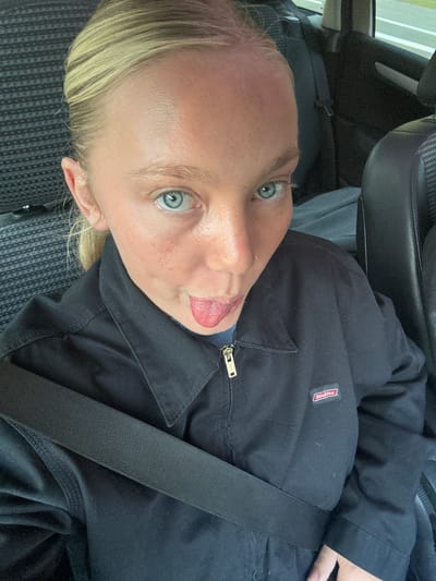 A person is in a car with a seatbelt fastened, sticking their tongue out at the camera. They are wearing a black zip-up jacket with a "Dickies" logo on the left breast. The background shows the car's interior, including the seat and window. The location is unknown and cannot be determined from the image. The location of the city of Serris, France is not discernible from the provided image.