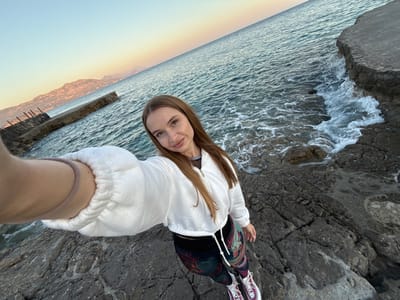 A young woman is depicted in a selfie, standing on a rugged, dark grey rocky coastline. The camera is held by her right arm, angled downwards to capture her full body and the surrounding landscape. She has long, straight light brown hair and is wearing a white, textured hooded sweatshirt with a zipper, layered over a black top. Her legs are clad in distinctively patterned leggings featuring intricate designs in green, red, and orange against a dark background. She wears athletic sneakers with white laces and pinkish-purple accents.
The woman is positioned close to the water's edge, with foamy waves of the sea gently crashing against the rocks to her left. The vast expanse of the ocean dominates the middle ground, its surface rippling under a clear sky. In the distance, a hazy mountain range stretches across the horizon, bathed in the warm, soft glow of either sunrise or sunset, creating a serene gradient from light blue to pale orange and yellow in the sky. To the right, a dark, linear concrete or wooden structure, possibly a dilapidated pier, extends into the water. The woman looks directly at the camera with a slight, gentle smile, conveying a sense of calm. The scene captures a tranquil coastal environment in Seoca, Montenegro, at a picturesque time of day. No visible text is present in the image.