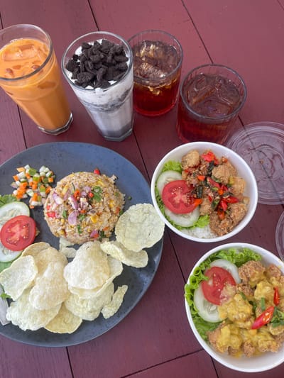 On a reddish-brown wood-grain surface, an arrangement of food and beverages is presented. From left to right, a glass of iced tea, a glass of Oreo milkshake, and a glass of Thai iced tea are visible. Two white bowls contain what appears to be rice topped with fried chicken and garnished with vegetables. A black plate features fried rice, diced vegetables, a slice of tomato, cucumber, lettuce, and a side of chips. Two glasses of iced tea with visible ice cubes complete the composition. The setting suggests a restaurant or cafe, with the city of Ngasinan, Indonesia, as the location.