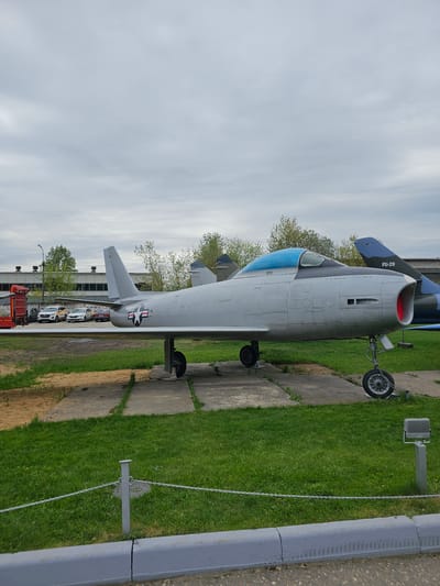 The image shows a North American F-86 Sabre fighter jet displayed outdoors. The jet is painted in a light gray color with blue accents on the canopy and tail. The fuselage bears the US national insignia. The aircraft is parked on a concrete surface alongside a grassy area. A metal chain fence runs parallel to the jet. In the background, buildings and several parked cars can be seen. The sky is overcast.
