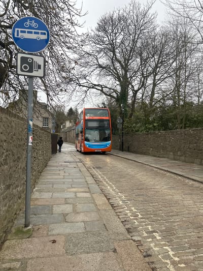 A slightly tilted, street-level shot captures a narrow, cobblestone street in Aberdeen City, United Kingdom. An orange and blue double-decker bus advances down the street toward the camera. A lone figure is walking in the same direction as the bus ahead of the bus. The street is flanked by stone walls on both sides. In the lower left corner, a sign attached to a metal pole shows a blue circular sign depicting a bus with a bicycle on top and a black and white rectangular sign featuring a camera. Bare trees frame the scene against a gray sky.