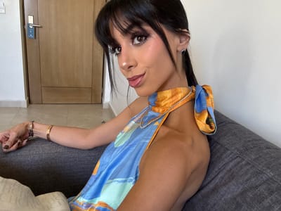 A woman with long dark hair and brown eyes is lying on a gray couch, wearing a colorful blue and orange top. She has multiple gold bracelets on her right wrist. A wooden door is visible in the background. The woman gazes directly at the viewer. The photo is oriented sideways.
