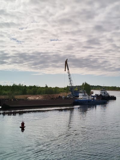 The image depicts a river scene at Ozernoe Ust'e, Russia. A long, dark barge filled with sand or gravel is docked alongside a blue and white tugboat with a large crane. A second, similar tugboat is positioned behind the first. A red buoy is visible in the foreground of the water, and a tree line stretches along the horizon. The sky is overcast with mottled clouds.