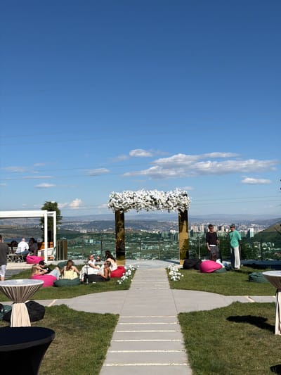 This media file depicts an outdoor gathering, likely a wedding ceremony or reception, set against a scenic backdrop overlooking a city. The scene is bathed in bright daylight, suggesting a mid-morning or afternoon time. The sky is a clear, vibrant blue with a few wispy clouds, indicating pleasant weather.

Several individuals are present, some seated on the grass and others standing. They appear to be guests or participants in the event. One prominent object is a tall, gold-colored structure adorned with white flowers, serving as a decorative element or possibly an archway. Other decorative elements include cushioned seating on the grass and draped fabrics. The ground is a mixture of lush green grass and a paved pathway made of light-colored stones. In the background, a cityscape unfolds, characterized by buildings and verdant hills, hinting at a location like Lisi, Georgia.

The overall mood of the scene appears relaxed and festive. People are casually dressed, and their postures suggest they are observing or engaged in conversation. The presence of celebratory decorations and the gathering of people strongly suggest an event, possibly a wedding, given the floral arrangements and the set-up. The time of day is clearly daytime, with strong shadows indicating the sun is high in the sky. There is no visible text in the media file.