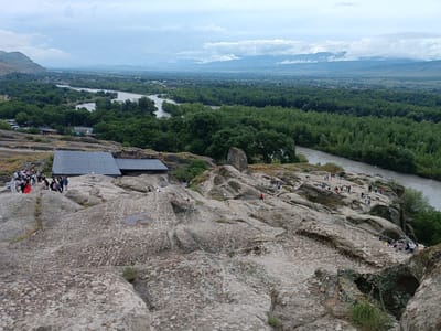 This is a wide-angle, high-angle, eye-level shot of the ancient cave city of Uplistsikhe in Georgia. The foreground is dominated by rough, weathered, beige-colored rocks with patches of lichen. In the middle ground, a group of tourists is visible on the rocks, some walking and others standing. To the left of the tourists is a dark grey, gabled roof structure, possibly a visitor center or museum.
Beyond the rocky terrain, a wide, muddy brown river flows, bordered by a dense, dark green forest. The forest stretches into the distance, where a hazy landscape of hills and mountains can be seen under a cloudy, overcast sky. The overall impression is of a historical site integrated into a natural landscape.