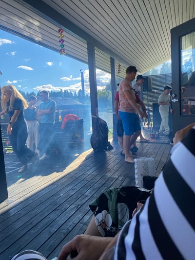 In Joensuu, Finland, a group of people are gathered on a wooden deck under a covered patio. The scene appears to be a social gathering, possibly a party or barbecue, given the presence of smoke and the relaxed attire of the individuals.
In the foreground, a person wearing a black and white striped shirt is partially visible, with their arm resting on a black and green floral-patterned fabric.
On the deck, several individuals are visible. A woman with blonde hair, wearing a dark blue outfit, is dancing on the left side of the image. To her right, a woman in a light-colored outfit stands next to a man in a light blue polo shirt who has his arms crossed. Further back, a man in a coral-patterned shirt and blue shorts is talking with other individuals, one of whom is wearing a white t-shirt and a black baseball cap. Another man in a light-colored t-shirt and pants is standing near a doorway, holding a white object.
The environment includes a wooden deck, a covered patio with a slatted ceiling, and a backdrop of trees and houses under a bright blue sky with scattered clouds. Decorative colorful paper ornaments are hanging from the ceiling of the patio. There are also speakers and lighting equipment present, suggesting music might be playing. A black trash bag is visible on the right side of the deck. Smoke is present, indicating a possible barbecue or a fog machine.