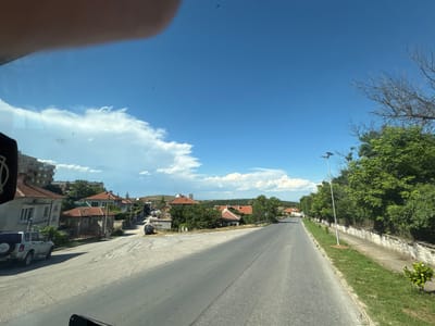 The image depicts a road winding through a residential area in Topolovgrad, Bulgaria. The scene is dominated by a clear blue sky with scattered white clouds. On the left side of the road, several houses with red tile roofs are visible, along with a silver SUV parked on the side. A multi-story building can be seen further down the road. The right side of the road features a sidewalk bordered by a stone wall and lush green trees. A lamppost with a solar panel stands on the right side of the sidewalk. The road appears to be made of asphalt and shows signs of wear. The overall impression is of a sun-drenched day in a quiet, rural town.