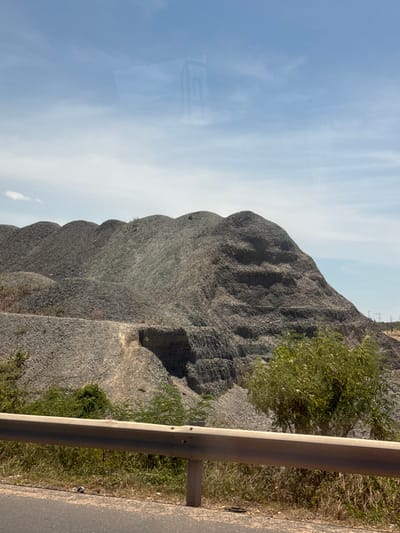 The image displays a large, grey, rocky mound, likely a pile of excavated earth or construction debris, under a bright, partly cloudy sky. The mound has a textured, layered appearance with visible undulations and shadows, suggesting a significant accumulation of material. To the right of the mound, there is a concrete barrier, possibly a guardrail, running parallel to a paved road or pathway. Sparse green vegetation, including bushes and small trees, grows in patches around the mound and along the side of the barrier. The scene appears to be outdoors, during daylight, with the sun casting shadows that indicate mid-morning or afternoon. The overall impression is one of an industrial or construction site, or perhaps a natural geological formation altered by human activity. There are no people visible in the image. A faint, rectangular symbol, possibly a logo or text, is reflected on the surface of the mound, to the left of the frame, appearing as a pale shape against the blue sky. The environment suggests a dry, possibly warm climate. The image is oriented vertically.