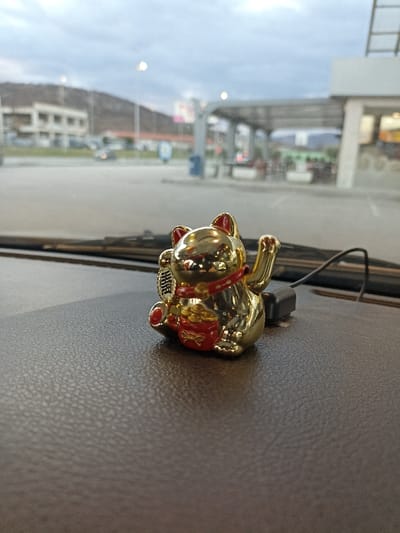 The image captures a close-up view of a golden Maneki-neko (lucky cat) figurine placed on what appears to be a car dashboard. The figurine is shiny gold with red accents on its ears, collar, a small bag of coins, and parts of its raised paw. The cat has a smiling expression. In its left paw, it holds a gold coin or plaque with visible black Chinese characters. Its right paw is raised in the traditional beckoning gesture. Behind the figurine, a black cable is connected to a small black rectangular object, suggesting it might power the cat's waving paw.

The dashboard surface is dark brown or black with a distinct textured, possibly leather-like, pattern.

Through the car's windshield, an out-of-focus outdoor scene is visible. The sky is overcast and appears cloudy, suggesting a gray day or possibly early morning/late afternoon. In the distant background on the left, a light-colored building with multiple windows is visible, situated against a blurred, dark brown hill or mountain range. Further to the right, structures resembling a gas station or car wash facility are apparent, with a large, light-colored canopy supported by pillars. Several indistinct vehicles are also visible in the blurred mid-ground, including one car in motion or parked in the middle-left of the background. The overall setting appears to be a parking lot or a service area.