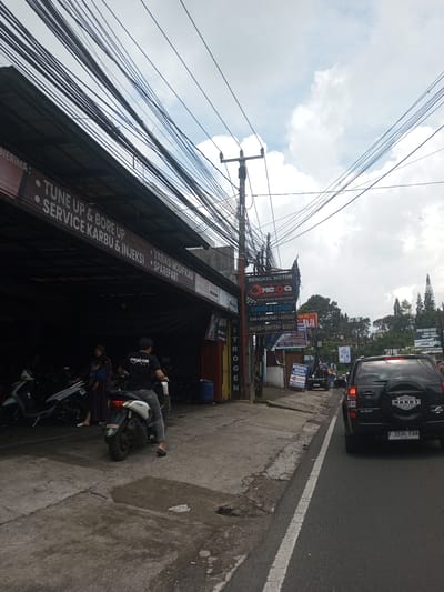 The image shows a street scene with a motorcycle repair shop on the left and a black SUV in the street on the right. The repair shop, "Bengkel Motor," is open, with motorcycles visible inside. A man on a motorcycle is facing the road, and a woman holding a baby is standing near the shop. Numerous electrical wires are overhead. Various signs are posted near the shop advertising services and products. The SUV has a spare tire on the back and a license plate that reads "F 1534 FDA." The location is likely in Indonesia based on the language on the signs. The city is NOT Ban Phouviengnoy, Laos.