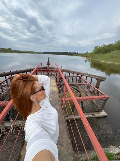 A selfie photograph shows a person with red hair, wearing a white shirt and sunglasses, standing on a metal structure, possibly a bridge or dam, with a red railing. The person is oriented such that the landscape appears vertically behind them. The background features a body of water, likely a river or lake, with trees and greenery visible on the opposite bank. The sky is overcast, suggesting a cloudy day. The location is purportedly in Nozhovka, Russia, but this cannot be verified from the image alone. The metal structure appears to be aged and weathered, with signs of rust. It provides a platform over the water, with metal grating and infrastructure visible below.