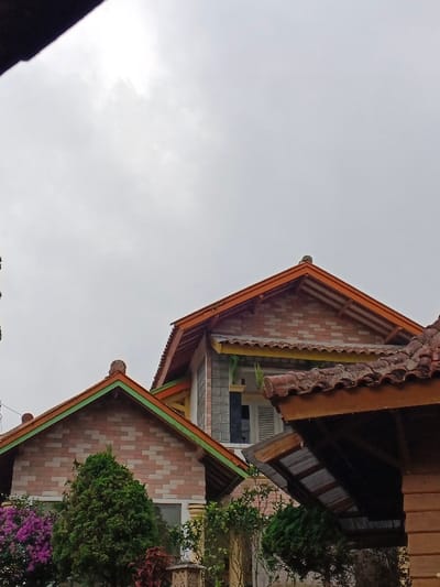The image depicts an exterior view of residential structures and vegetation in Kyauk Hsoke, Myanmar, under an overcast sky. The upper two-thirds of the frame are dominated by a uniform, light grey, cloudy sky.
In the foreground, a dark, angular object partially obscures the upper-left corner of the frame. Below this, on the left, a light-colored brick house with a green-trimmed gable roof is visible, partially obscured by a dense green conical shrub and purple flowering bougainvillea. To the right of this, a more prominent two-story structure features an orange-brown-trimmed gable roof above a brick facade. Below its main gable, a secondary corrugated roof with yellow and green trim is present. A vertical window with glass block segments and a white louvered shutter is visible on the upper level of this structure. Further to the right, in the lower-right foreground, a dark brown wooden structure with a weathered, reddish-brown tiled roof is partially visible, featuring a corrugated metal overhang beneath it. No individuals are present in the scene. The environment appears residential with significant natural foliage.