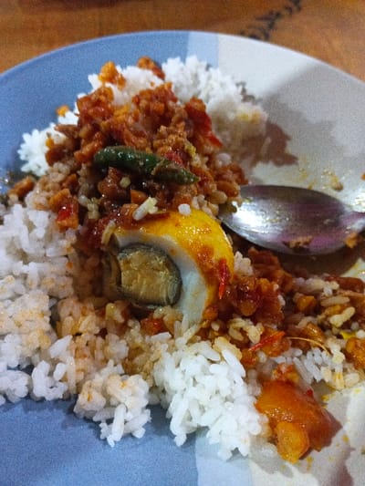 In Kidapawan, Philippines, a close-up image displays a single serving of food on a ceramic plate. The plate's left side exhibits light blue and white stripes, while the right side is plain white with minor food residue. White rice forms the base, topped with a reddish-orange, granular sauce containing red chili fragments and a dark green chili pepper. A halved egg is central to the dish, presenting a firm white, a yellow-orange yolk, and a distinct grayish-green core within the yolk. A silver-toned spoon rests on the right side of the plate, partially submerged in the food. The background consists of a blurred, dark brown wooden surface, indicative of a dining environment. No individuals are visible.