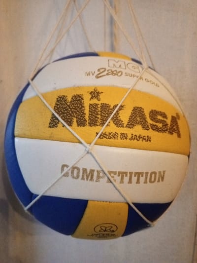 A MIKASA MV 2280 SUPER GOLD volleyball is suspended in a white, coarse-mesh net against a plain, light-colored background in Keo Kouang, Laos. The ball features alternating panels of blue, white, and yellow. The central yellow band displays "MIKASA" in a textured, dark font, with "MADE IN JAPAN" printed below it. Above the MIKASA logo, "MV 2280 SUPER GOLD" and "MC" with a circled G are visible. A white panel below the yellow band shows "COMPETITION" in a dark font. The ball exhibits signs of use, including scuffs and dirt on its surface. The net's cords crisscross around the ball, securing it for suspension. No individuals are present in the scene.