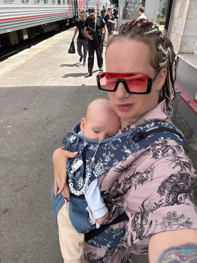A woman with braided hair and large pink-tinted sunglasses carries a baby in a sling carrier at a train station in Grushevskaya, Russia. She wears a light pink blouse with a dark floral and animal print. The baby, facing the woman, is dressed in a light blue onesie and is secured in a dark blue sling with a patterned overlay. A beaded pacifier clip hangs from the sling. In the background, a train is visible on the tracks, and several people are walking on the platform. Some individuals appear to be in uniform, possibly security personnel, with one man in a dark uniform carrying a black plastic bag. The ground is paved with asphalt, and the lighting suggests it is daytime.