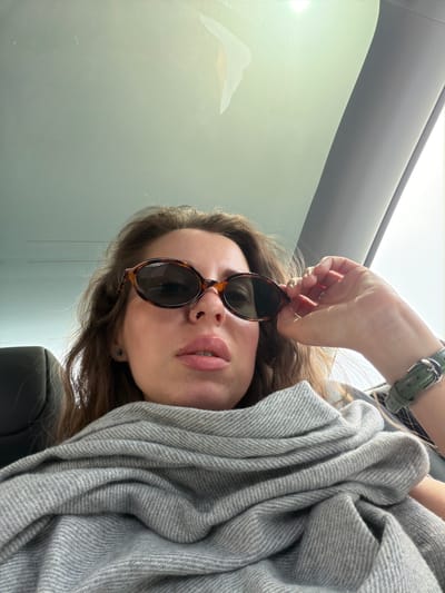 A woman with brown curly hair, wearing a gray herringbone shawl and brown sunglasses, holds the sunglasses near her face. A nose ring is visible. She is in the passenger seat of a car. The location is likely within the Menashe Regional Council, Israel, though no specific landmarks are present.