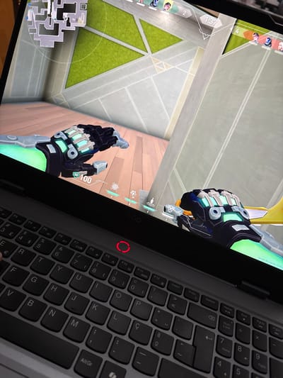 The image captures a first-person perspective video game being played on a black laptop, angled from the side to show both the screen and part of the keyboard.

On the left side, the laptop screen displays a tactical first-person shooter game, likely Valorant, given the distinctive user interface and character art style. The in-game view shows robotic, cybernetic hands, suggesting a character with advanced technology. The environment rendered in the game features a stylized, futuristic setting with light-colored, geometric concrete or metallic structures and patches of bright green, possibly grassy areas.

Various game UI elements are visible:
-   A minimap is present in the bottom-left corner, showing a detailed layout of the game area.
-   On the left edge, a vertical stack of player portraits with numbers (e.g., "2", "4") indicates team status or scores, with "1:37" displayed as a timer.
-   In the bottom-right of the screen, the character's health is indicated as "100" with "50" next to it, likely representing armor or shield. Several ability icons are arranged above this display.
-   In the top-right corner, the text "Versão do cliente: 12.02" ("Client version: 12.02") is visible.

The right side of the image features the laptop's keyboard. It is dark grey with black keys and has a European layout, indicated by symbols like '€' and 'ç'. Visible keys include a range of function keys (F4 through F12), special keys like Insert, PrtSc, Delete, Home, End, PgUp, PgDn, and Num Lock, along with standard alphanumeric and symbol keys. A small red circular indicator or power button with a red outline is visible next to a "Legion" brand logo above the keyboard, near the screen hinge.