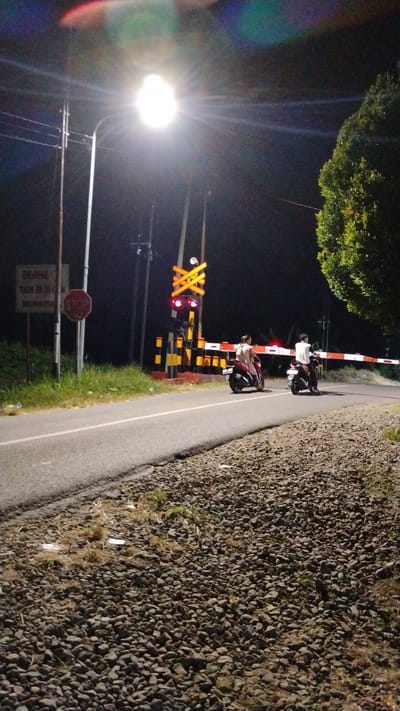 The image depicts a nighttime scene at a railway crossing in Tulungrejo, Indonesia. A bright street lamp illuminates the area, casting lens flares and creating a halo effect. Two individuals on motorcycles are stopped at the railway crossing. The crossing has red and yellow striped barriers that are lowered, indicating an approaching train. Red lights are flashing on the crossing signal. A "STOP" sign is visible on a post to the left, along with another sign with Indonesian text. The road is paved, and the area to the side of the road is covered in gravel and dry grass. A large, leafy tree is visible on the right side of the frame. The overall atmosphere suggests a moment of waiting at a railway crossing at night.