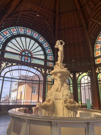 The image depicts the interior of the Palais Lumière in Évian-les-Bains, France, a former thermal spa now serving as a cultural center and exhibition space. Dominating the foreground is a large, ornate, off-white stone fountain sculpture. The fountain features a central figure of a nude woman with her arms raised, standing atop a tiered base adorned with cherubs and architectural motifs, including what appear to be carved shells or urns.
Behind the fountain, a series of tall, arched windows with stained-glass accents allow natural light to filter into the space. The stained glass features intricate patterns of floral designs in shades of red, orange, purple, and blue. The windows look out onto a street scene with buildings, including a prominent orange building with balconies and windows, under a bright sky.
The interior structure is characterized by exposed wooden beams forming a vaulted ceiling, characteristic of Belle Époque architecture. Wooden decorative elements and frames are visible throughout the space, surrounding the windows and supporting the ceiling.
In the middle ground, a curved, light-colored counter or railing encloses the fountain area. A red rope barrier is visible along this counter. A woman with short dark hair, wearing a light-colored top, stands behind the counter, partially obscured by the fountain sculpture. In the background, through the windows, several empty wicker chairs are visible, suggesting seating areas.
The overall impression is one of grandeur and historical significance, showcasing the architectural beauty of the Palais Lumière and its centerpiece fountain. The lighting is bright, with natural light coming from the windows, highlighting the details of the sculpture and the stained glass.