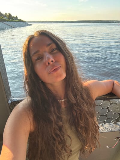 The image is a selfie taken by a young woman with long, brown hair. She is wearing a light olive-colored top and a thin, light-colored necklace. The setting appears to be outdoors, near a body of water, likely a river or lake, given the presence of the shoreline with trees and buildings in the distance. The water has ripples, suggesting a gentle breeze. The sun is low in the sky, casting a warm, golden light, indicating it is either sunrise or sunset. The woman's face is turned towards the camera, and she has a relaxed, content expression. She is leaning on a dark-colored railing, which has a decorative pattern of circular metallic accents. The overall mood of the image is peaceful and serene, capturing a moment of quiet enjoyment in a natural setting. The location is identified as Verhnij Uslon, Russia. There is no visible text in the image.