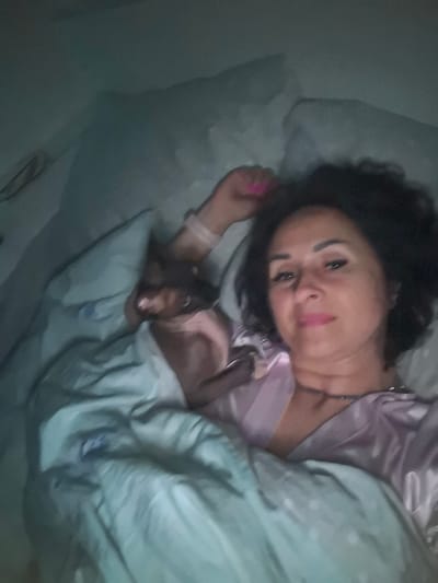 The image depicts a woman lying in bed, partially covered by a light blue comforter and a pink satin robe. Her dark, curly hair is framing her face, and she is looking towards the camera with pink lipstick on. A small, light-colored animal, possibly a cat, is nestled beside her under the covers. The room appears dimly lit, suggesting it is nighttime or early morning. The location of Teykovo, Russia, is not depicted or suggested by any visual elements in the image.