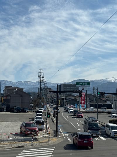 A street scene with traffic is set against a mountainous backdrop with a partly cloudy sky. Utility poles with wires line the road. A large green sign, partially obscured, stands along the street. The traffic light is green. Cars line up at a crosswalk. Buildings are visible on both sides of the street. The image is oriented vertically.
