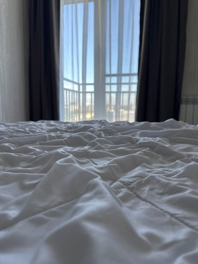 The image depicts a low-angle view of a rumpled white duvet on a bed, facing a large window with sheer white curtains and dark brown drapes. The window offers a blurred view of a cityscape and a balcony railing. The scene is indoors, likely a bedroom, with a radiator visible on the right side of the frame. The lighting suggests it is daytime. The provided location, Челюскинский, Russia, suggests this bedroom is situated in that urban area.