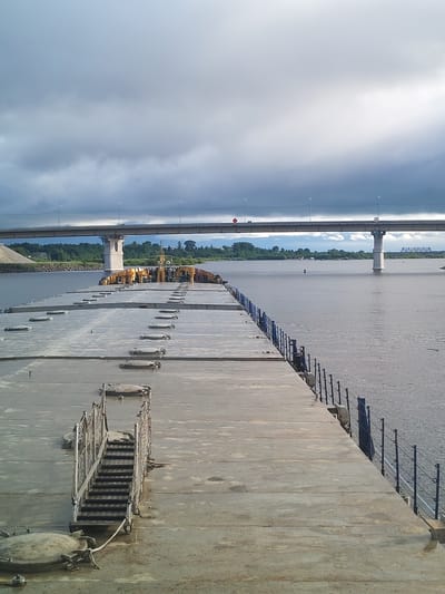A low-angle, wide shot captures a gray river scene in what is likely Нифантово, Russia, under an overcast sky. A long, flat, concrete barge stretches from the lower center to the middle of the frame, its surface marked with numerous rectangular hatches. On the right side of the barge, a dark blue railing runs along the edge. At the forefront, a metal staircase leads up to the barge's surface, adding a sense of scale and detail.
In the background, a concrete bridge spans the river, connecting the landscape. Green vegetation lines the riverbank in the distance. The water surface is calm, reflecting the muted tones of the sky above. The overcast sky dominates the upper portion of the image, suggesting a potentially somber or cool weather condition. No people are visible in the shot.