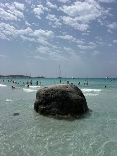 The image depicts a lively beach scene in Crabonaxa, Villasimius, Italy. The foreground is dominated by a large, dark boulder partially submerged in crystal-clear turquoise water. The water is shallow, with gentle waves lapping at the shore and revealing the sandy seabed. In the middle ground and background, numerous people are enjoying the water, swimming, and wading. Some are standing, others are floating, and a few are lying down in the water. The sea is dotted with various sailboats and a motorboat, indicating a popular nautical area. In the distance, the coastline is visible with hills and a structure that appears to be a tower. The sky above is a bright blue, adorned with scattered white, fluffy clouds, suggesting a sunny day.