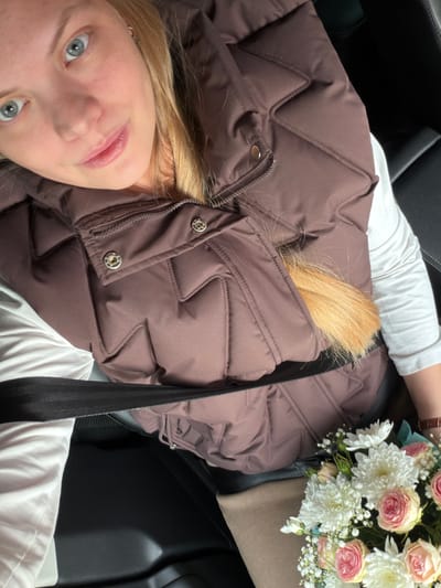 A young woman is captured in a close-up, low-angle shot, seemingly taken from inside a car in Uspenskii, Russia. She has blonde hair and light blue eyes, looking directly at the camera with a soft, pleasant expression. She is wearing a white long-sleeved top underneath a dark brown, quilted puffer vest. A black seatbelt crosses diagonally across her chest and shoulder. Part of her blonde hair spills over the top of the vest.

Next to her, on what appears to be a car seat or armrest, rests a bouquet of fresh flowers. The bouquet includes light pink roses, white chrysanthemums, and small white baby's breath, wrapped in beige paper with a hint of light blue ribbon or paper visible. A watch with a brown leather strap is visible on her left wrist, near the flowers. The car's interior features dark upholstery, visible as black seats around her. The image conveys a casual, personal moment, possibly on a cool day given the puffer vest. No text is visible in the image.