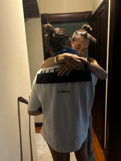 Two women embrace indoors. The woman facing forward wears a white t-shirt with black vertical stripes and the word "LONDON" printed. The other woman has her hair in a bun and wears a blue top. A dark paw and leg of a dog are visible near the bottom of the frame. The scene is warmly lit, with a wooden door and window frame in the background.