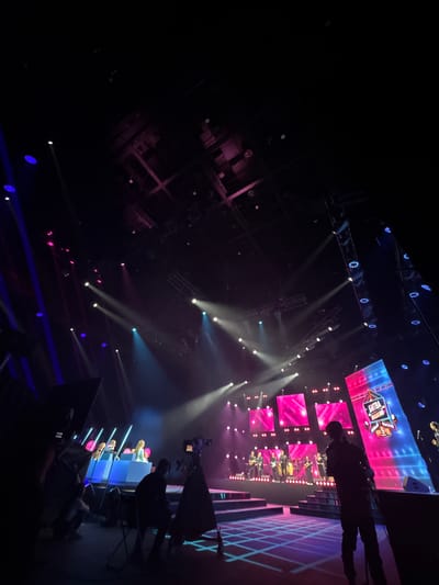 The image shows a stage in a dimly lit venue, likely a concert hall or television studio, in Nemchinovka, Russia. The stage is illuminated by vibrant pink and blue lights, with several spotlights casting beams across the ceiling and down onto the stage floor, which has a grid pattern of light.

In the background, a band is performing on a raised platform, with musical instruments visible. A large screen behind the band displays the text "Битва Поколений" (Battle of Generations) and other graphics.

In the foreground, several people are present. To the left, a group of individuals are seated behind a blue barrier, possibly judges or contestants. To the right, a person in silhouette is standing in front of a camera, suggesting a filming or broadcast event. Other figures are also visible in the shadows, some appearing to be part of the production crew.

The overall atmosphere is one of a lively performance or event, with a focus on visual spectacle provided by the lighting and stage design.