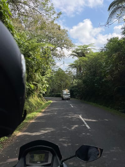 A first-person perspective shot from a motorcycle looking forward along a paved road surrounded by lush green vegetation and trees. The road is empty except for a white truck in the distance, driving in the same direction. The sky is partly cloudy with a bright blue sky. The motorcycle's dashboard is visible in the foreground, displaying a speed of 13 km/h. The scene appears to be a rural area with dense foliage and winding road, consistent with locations in Pupuan, Indonesia.