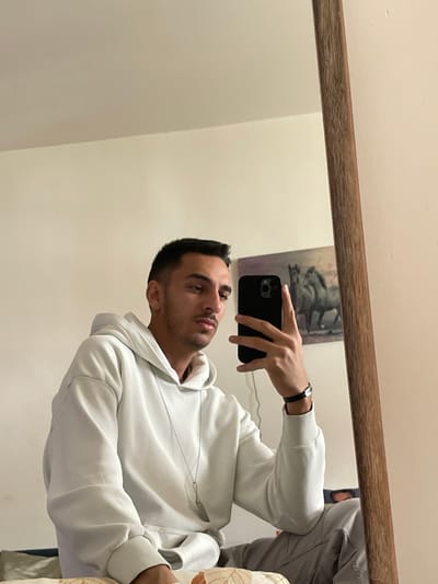 The scene takes place in an indoor residential setting, specified as being within the Neapoli Municipal Unit, Greece. A young adult male is captured in a mirror selfie, reflecting his upper body and head. He has short, dark hair and subtle facial hair, looking directly towards the smartphone in his right hand. He is wearing a light-colored, possibly off-white, hooded sweatshirt with a long-sleeved design, and a silver-toned necklace with an elongated pendant hangs around his neck. A dark wristband or watch is visible on his left wrist. The black smartphone, with a visible multi-lens camera module, is held vertically. The background, as seen in the mirror, features a plain light-colored wall. A framed artwork depicting two horses in muted tones is mounted on the wall to the subject's right. The mirror itself has a light brown, wood-grained frame, partially visible on the right side of the image. A section of a patterned fabric, possibly part of a bed or couch, is visible in the lower-left corner, suggesting the individual is seated.