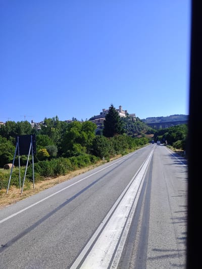 The image depicts a view from a vehicle traveling on a road in Spoleto, Italy. In the distance, atop a hill, stands a prominent stone complex, possibly a fortress or castle, with multiple buildings and towers. To the right of the complex, a large, old stone aqueduct arches across a valley. The road is paved and has white lane markings. Green trees and vegetation line the sides of the road, with a few buildings visible in the background on the hillside. On the left side of the road, a black sign on a metal stand is positioned. The sky is clear and blue. There are two vehicles visible on the road ahead, a dark truck and a white van. The overall scene suggests a sunny day in a rural or semi-rural Italian landscape.