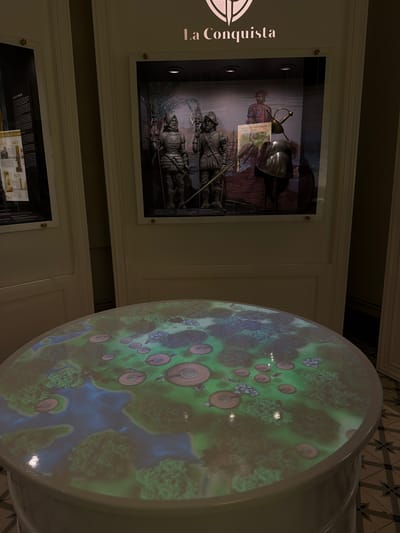 The image shows a museum exhibit with historical and geographical content. On the left, a framed display labeled "La Conquista" presents a depiction of the Spanish conquest. The exhibit includes images of conquistadors, arms, and what appears to be historical documentation. To the right is a large, rounded surface displaying a projection of a map featuring blue areas representing water, green areas representing land, and circular brown features that could represent settlements or other geographical features. The projection is likely intended to provide a spatial or environmental context related to the theme of "La Conquista." The scene seems to be set within a museum or similar educational environment with patterned flooring visible in the background. Without additional context, a specific location in Tampico, Mexico cannot be identified.