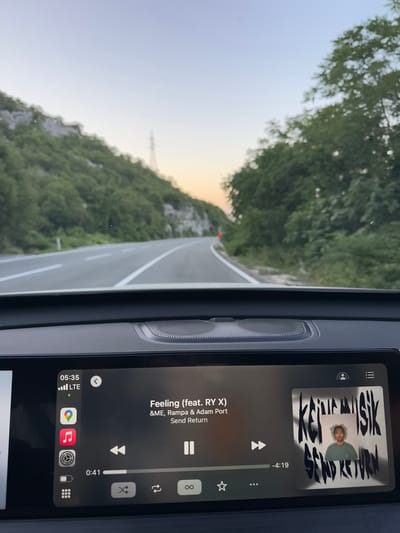 The image shows a dashboard view from inside a vehicle driving on a scenic road. The road curves to the left, with steep, tree-covered hills on both sides. The sky is clear with a pale orange hue near the horizon, suggesting it is either sunrise or sunset. A power line tower is visible on the left hill. The car's infotainment system is displayed on the dashboard screen, showing song information for "Feeling (feat. RY X)" by &ME, Rampa & Adam Port from the album "Send Return". The time displayed is 05:35 and the LTE signal is strong. There are icons for navigation (Google Maps), music (Apple Music), settings, and other apps on the left side of the screen. The song is currently playing, with the progress bar indicating 0:41 out of 4:19. The album art for "Send Return" is displayed on the right side of the screen. The car is in motion, as indicated by the road ahead and the dynamic nature of the scene. The location is identified as Gornja Brova, Montenegro, a mountainous region known for its natural beauty.