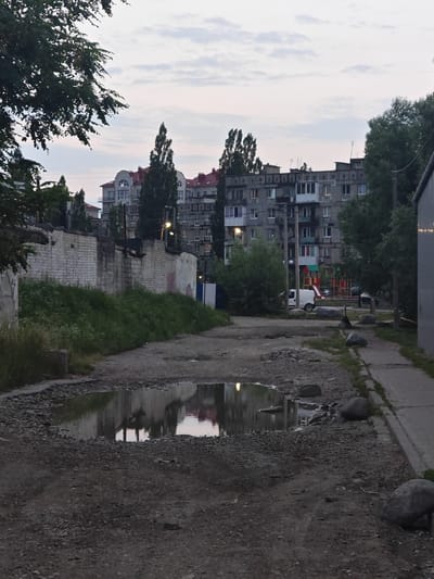 The image depicts a street scene in Vasilkovo, Russia, characterized by a rough, unpaved road in the foreground with a large puddle reflecting the surrounding buildings and sky. A sidewalk runs along the right side of the frame, bordered by grass and large stones. In the background, multi-story apartment buildings, typical of Soviet-era architecture, are visible, some with red-tiled roofs. A playground with slides and swings is situated between the buildings. A white van is parked near the playground. A low, weathered brick wall with graffiti runs along the left side of the road, with overgrown green vegetation behind it. Trees are scattered throughout the scene, with dense foliage on the left and right edges of the image. The sky is overcast, suggesting it is either early morning or late evening. There are no people actively engaged in any discernible actions.