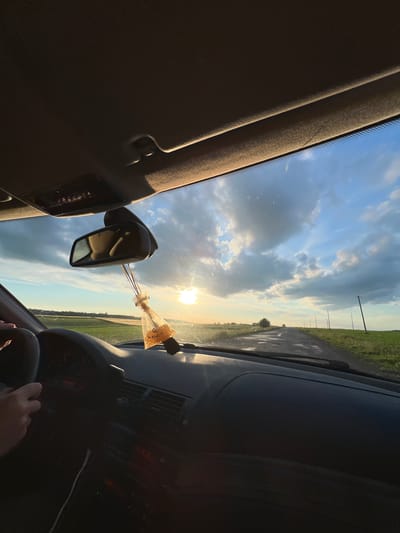 The image presents a view from the interior of a vehicle, looking forward through the windshield in Skomorokhy, Ukraine. The scene depicts a rural road and landscape at sunset. Inside the vehicle, the dark headliner is visible at the top, with an overhead console and rearview mirror centrally positioned. A "DR. MARCUS" brand air freshener, a mesh bag containing yellow beads, hangs from the rearview mirror. The dark dashboard, including parts of the instrument cluster and air vents, occupies the lower half of the frame. A person's hands are visible on the steering wheel on the left, indicating a driver. Outside, a narrow, unpaved or damaged asphalt road with visible wet patches extends towards the horizon, bordered by expansive green fields. The setting sun is low and bright on the horizon, directly ahead, causing a strong glare on the windshield. The sky above features scattered white and gray clouds against a blue backdrop. Multiple utility poles with wires are visible along the right side of the road, receding into the distance.