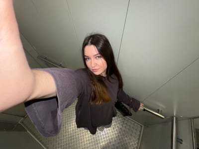In Shirkovo, Russia, a young woman with long, dark hair takes a selfie from a high angle. She is wearing a dark purple, long-sleeved shirt and denim shorts. Her right arm is extended to take the photo. She is standing on a metal grate floor, with a silver railing visible to her right. The background consists of smooth, light-colored walls. The lighting appears to be artificial.