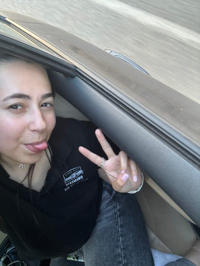 The image depicts a single individual, a young female, seated within the interior of a vehicle in Omarchevo, Bulgaria. She has dark hair, dark eyebrows, and light-colored skin. Her mouth is slightly open, and her tongue is extended in a playful gesture. Her left hand (viewer's right) is raised, forming a peace sign with her index and middle fingers. Her fingernails are painted a light lavender or pale pink. A ring is visible on her left ring finger, and a thin bracelet is on her left wrist. She wears a silver-toned necklace with a double cloud or interlocking heart pendant. The individual is dressed in a black collared shirt featuring a white logo that reads "SINCE 2024 BENZMANIA OUR RACING TEAM" alongside a stylized race car graphic. She is also wearing dark wash jeans. The car's interior shows dark grey textured plastic, likely a door panel or dashboard, and lighter tan or brown upholstery. The exterior view through the car window is blurred with horizontal streaks, suggesting the vehicle is in motion on a road.