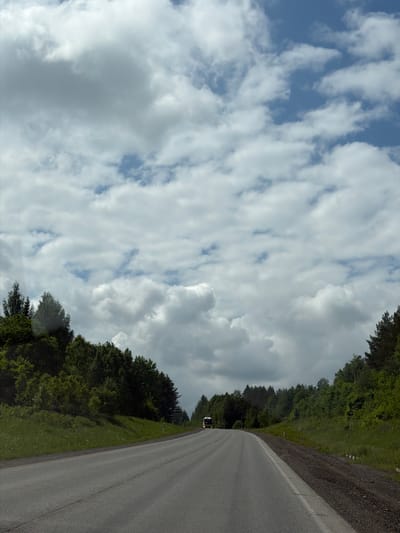 The image displays a rural road scene, captured from a slightly elevated perspective, with the original image appearing rotated 90 degrees clockwise. When viewed upright, the scene features a two-lane asphalt road curving gently into the distance on the right side of the frame, bordered by a dirt and gravel shoulder. A single white line is visible on the asphalt, likely marking the edge of the lane or shoulder.
To the left of the road, dense green trees and foliage form a thick forest that stretches towards the horizon. The trees appear lush, indicating a temperate climate. A small, light-colored vehicle, possibly a white van or truck, is visible on the road in the mid-ground, heading away from the viewer.
Above the tree line and road, the sky is filled with a mix of cumulus and stratocumulus clouds. Patches of clear blue sky are visible between the clouds, suggesting a partly cloudy day rather than an overcast one. The lighting indicates it is daytime. No specific time of day can be determined beyond daylight hours. There is no visible text or identifiable people in the image. The overall impression is one of a peaceful, natural landscape along a country road in Башкултаево, Russia.
