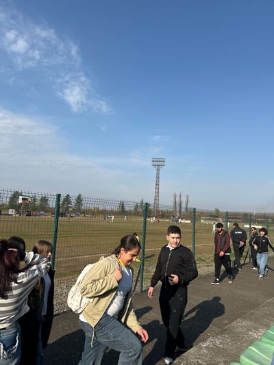 The scene depicts an outdoor football stadium or training facility in ქალაქი ლანჩხუთი, Georgia, on a clear, sunny day with a blue sky and sparse white clouds. A green chain-link fence separates a paved pathway in the foreground from a football pitch in the mid-ground. On the pitch, multiple individuals in white and blue jerseys are engaged in a football match or practice. A tall, reddish-brown metal floodlight tower stands prominently in the mid-ground to the right of the field's center, and a smaller, elevated structure bearing a 'FIFA' logo is visible on the far left side of the field. Trees, some bare, line the perimeter of the facility.
In the immediate foreground, a group of four young individuals walks along the paved path. A female in a tan jacket, white top, and blue jeans, carrying a white backpack on her left shoulder, is stepping down and looking towards the ground with a slight smile. To her right, a male in a black bomber jacket and black track pants walks forward, holding a dark object in his left hand. Two other young females are partially visible on the far left. Further along the path in the mid-ground, a group of approximately 5-7 males walks from right to left. One male wears a reddish-brown jacket and dark pants, and another behind him wears a black hoodie with 'JACOMOMO' branding. Green stadium seating is partially visible in the lower right corner of the frame.