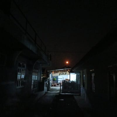 This is a nighttime exterior shot of a building in Cigadung, Indonesia, with a sign reading "DAYTONA" in English and Japanese characters. The building appears to be a business, possibly a shop or garage, with a fenced-in area visible behind the sign. The moon is visible in the sky as a small, bright orb. The scene is dimly lit, with the primary light sources being from inside the building and the moon. The overall impression is one of a quiet, somewhat isolated location.