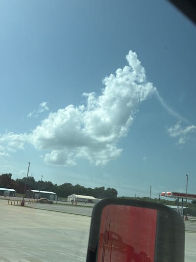 The image captures a wide-angle view of a truck's side mirror reflecting a bright red surface, likely the truck's body, against a backdrop of a suburban or rural landscape. The sky is a clear, pale blue with scattered cumulus clouds, one of which is particularly prominent and resembles a fantastical creature. In the distance, a gas station with red and white trim and a sign displaying a red circular logo is visible. Several low-lying buildings with white walls and green roofs are situated near the gas station, surrounded by lush green trees. A solitary gold-colored car is parked on a paved surface in front of one of the buildings. Several lampposts are also dotted across the scene. The image appears to be taken on a sunny day. The location is identified as Cardwell, United States.