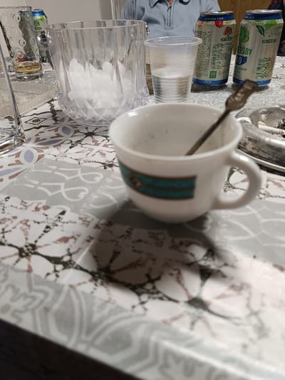 The image captures an indoor scene at a table in Izbeglii, Bulgaria, featuring beverages and an ashtray. In the foreground, a white ceramic demitasse cup with a teal blue and gold decorative band holds a dark metal spoon and contains residual liquid or sediment. This cup rests on a patterned surface displaying grey, white, and muted brown geometric and floral motifs.

Further back, a clear, faceted glass ice bucket is filled with ice cubes. To its immediate right, a clear plastic cup holds water. Adjacent and slightly behind the plastic cup, a circular metal ashtray contains several extinguished cigarette butts and ash. Two aluminum beverage cans are visible behind these items: one silver-colored Somersby branded can with red and green accents, labeled "NON ALCOHOLIC," and another silver-colored Somersby branded can with green accents.

In the upper-left background, a person wearing a light blue zippered top is partially visible, indicating their presence at the table. A dark-clad arm or shoulder of another individual is faintly discernible in the upper-right background. The scene suggests a casual social gathering involving drinks and smoking.